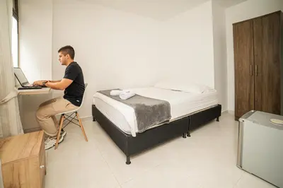 a man sitting on a chair next to a bed with a laptop at Descansa en Habitación Privada en Medellín C in Medellín